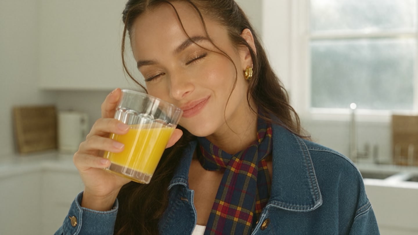 A woman in a denim jacket drinking a glass of orange juice in a bright kitchen. She is an AI-generated person.